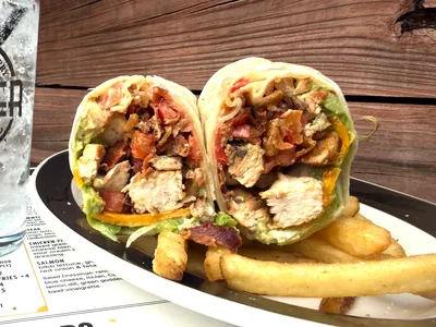 A grilled chicken wrap cut in half to show chicken, bacon, tomato, avocado, cheddar and lettuce inside, served with thick-cut fries on a plate beside a glass of ice water.