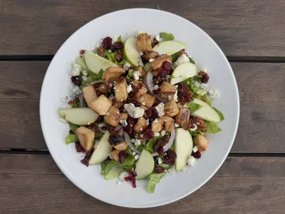 Overhead view of The Office salad on a white plate: grilled chicken, green apple slices, dried cranberries, crumbled blue cheese, bacon and red onion over mixed greens, on a wooden table.