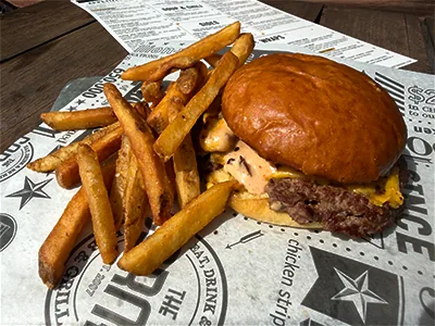 A smash burger with melted American cheese and creamy sauce on a toasted brioche bun, served with fries on Corner Pub & Grill branded paper.